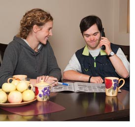 two people sitting at a table reading a newspaper and one is talking on the phone